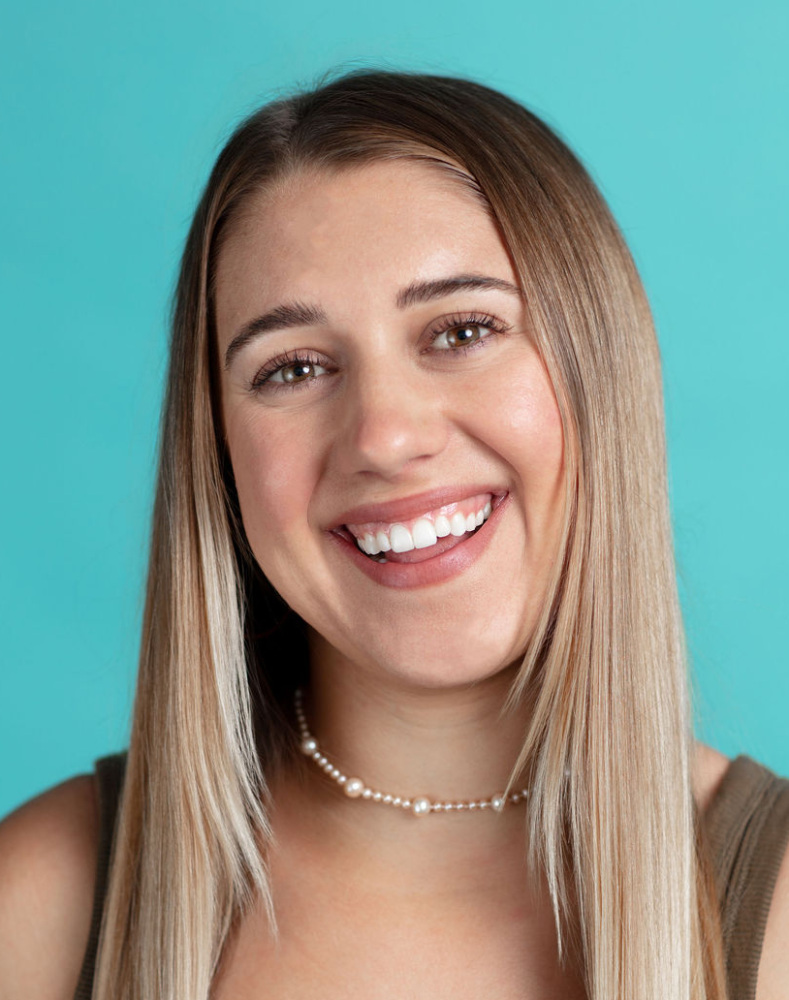 A young woman with straight, long blonde hair and a pearl necklace smiles at the camera against a teal background. She is wearing a sleeveless, ribbed olive top.